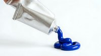 Blue paint pouring from silver tube onto white surface creating artistic puddle for creative projects and design work.