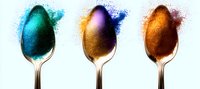 Colorful spoons with vibrant powder pigments in turquoise, purple, and orange creating artistic splash effects against white background, suitable for culinary or cosmetic designs.