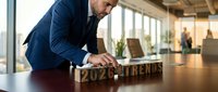 Middle-aged Caucasian businessman in navy suit arranging wooden letter blocks spelling 2024 TRENDS on conference table in modern office workspace.