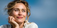 Young blonde woman with natural makeup smiling warmly while resting chin on hand against soft blue background for lifestyle and wellness content.