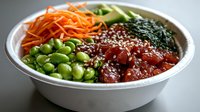 Fresh poke bowl with raw tuna, edamame beans, shredded carrots, cucumber, seaweed and sesame seeds served in white bowl on gray background.