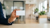 Professional holding blank white business card in modern office environment with plants and natural light for corporate branding and networking purposes.