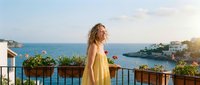 Young woman in yellow dress enjoying sunset on Mediterranean terrace overlooking azure sea and coastal village with blooming flowers.