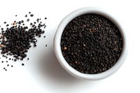 Black peppercorns in white ceramic bowl and scattered on white background, overhead view showing texture and natural spice ingredients for cooking and seasoning.