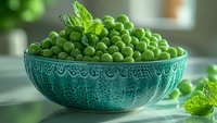 Fresh green peas in decorative turquoise ceramic bowl with mint leaves, natural window lighting creates soft shadows on kitchen counter, macro food photography.