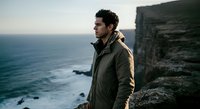 Young man with dark hair and olive complexion wearing olive jacket standing on rocky coastal cliff overlooking ocean, contemplative travel and adventure lifestyle portrait.