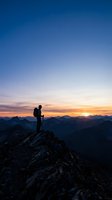 Silhouetted hiker with backpack standing on rocky mountain peak at sunset, gazing at golden sky and distant mountain ranges during golden hour adventure and outdoor exploration.