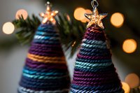 Handcrafted yarn Christmas trees with metallic star toppers against blurred holiday lights background, featuring stripes in purple, blue and gold tones.