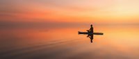 Silhouette of kayaker paddling on calm water during golden sunset with orange and pink sky colors reflecting on lake surface creating peaceful scene.