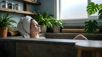 Young woman relaxing in bathtub with towel on head surrounded by green plants in modern bathroom spa setting for wellness and self-care routine.