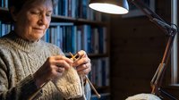 Senior woman knitting with needles and yarn in home library setting under desk lamp, engaged in handmade craft hobby.