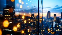 Modern cityscape at dusk with glowing bokeh lights in foreground, blurred urban skyline with skyscrapers against dramatic sunset sky creating atmospheric mood.