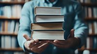 Stack of hardcover books held in hands against blurred library background, moody lighting creates atmospheric academic setting. Focus on textbooks with soft bokeh effect.