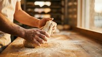 Baker hands kneading fresh dough on wooden counter in warm bakery kitchen. Traditional bread making process for artisan baking and culinary crafts.
