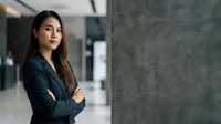 Confident Asian businesswoman in dark blazer standing with crossed arms in modern office corridor, professional portrait for corporate and business concepts.