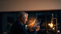 Senior businessman analyzing financial data on tablet in modern office with warm evening lighting and city view background.