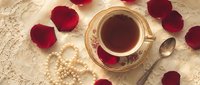 Elegant vintage teacup with black tea surrounded by red rose petals on delicate lace tablecloth, perfect for romantic breakfast or afternoon tea ceremony.