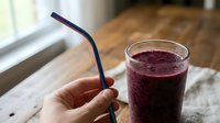 Person holding reusable straw while standing next to fresh berry smoothie on wooden table, promoting sustainable living and healthy drinking habits.