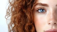 Young woman with natural red curly hair, bright blue eyes, and delicate freckles on fair skin shown in close-up portrait against light background.