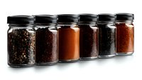 Glass jars filled with various ground spices and seasonings arranged in row on black background, selective focus on spice containers showing texture and color variety.