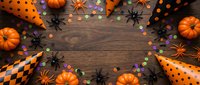Halloween party decorations with orange pumpkins, black spiders, polka dot paper, checkered patterns and colorful confetti on rustic wood background.