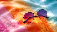 Stylish round sunglasses with purple-red gradient lenses on vibrant rainbow colored terry cloth beach towel against blurred summer background. For fashion and lifestyle content.