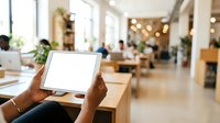 Person with dark skin tone holding tablet with blank white screen in modern office or coworking space with colleagues and natural light for app mockup or digital content display.