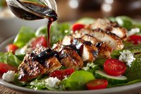 Fresh grilled chicken salad with balsamic glaze being poured over juicy meat slices, cherry tomatoes, feta cheese, and green lettuce leaves.