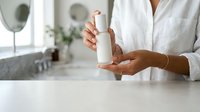 Woman with medium skin tone holding white cosmetic bottle in bathroom setting, showcasing skincare product for beauty and wellness marketing.