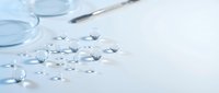 Transparent liquid droplets scattered on white surface with laboratory glassware and pipette, suitable for pharmaceutical, medical, and scientific research visuals.