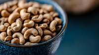 Raw cashew nuts in blue ceramic bowl on dark background, selective focus on golden brown nuts with natural texture and curves, food photography.