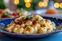 Homemade gnocchi pasta with crispy bacon bits and fresh herbs served on blue ceramic plate, soft bokeh lights in background creating cozy atmosphere.