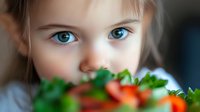 Close up portrait of young child with bright blue eyes looking at fresh strawberries and green leaves on plate, soft bokeh background, shallow depth of field.