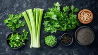 Fresh green celery stalks with parsley leaves and assorted spices in black bowls on dark stone background, overhead view of cooking ingredients layout.