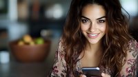 Young Hispanic woman with long wavy brown hair smiling while using smartphone at home. Wearing floral pattern dress in casual indoor setting with natural lighting.