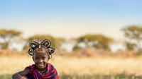 African woman wearing traditional beaded headdress and red fabric necklace smiles at camera in savanna landscape with acacia trees and golden grassland background.