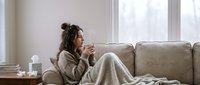 Young woman with long dark hair, South Asian ethnicity, sitting on beige sofa by bright window, holding warm beverage, wearing cozy cardigan and blanket, relaxed home setting, winter wellness.