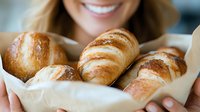 Woman with long brown hair and fair complexion holding fresh baked croissants and bread rolls in parchment paper, smiling at camera for bakery, food business, or culinary content.