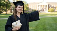 Young Caucasian female graduate in cap and gown pointing forward while holding education fund piggy bank on university campus during golden hour sunset.