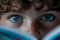 Young child with striking blue eyes and freckles reading book, close-up portrait showing curiosity and learning through literature and education.