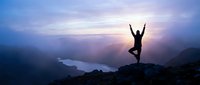 Woman practicing yoga tree pose on mountain peak at sunrise with lake valley below creating peaceful meditation scene for wellness lifestyle.