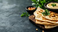 Fresh homemade naan flatbread served on wooden board with hummus and chickpea salad. Traditional Indian cuisine on dark stone background with copy space.