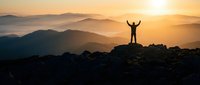 Silhouette of victorious person with raised arms standing on mountain peak at golden sunrise overlooking layered mountain ranges in misty landscape.