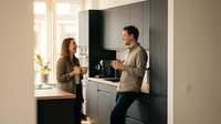 Young couple enjoying morning coffee together in modern minimalist kitchen with gray cabinets and natural light streaming through windows.