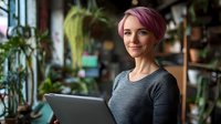Young Caucasian businesswoman with pink hair working on laptop in modern office with plants, smiling confidently at camera in coworking space environment.