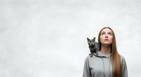 Young woman with long brown hair and fair complexion wearing gray hoodie, looking upward thoughtfully with black cat perched on shoulder against white textured wall background.