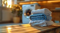 Fresh folded laundry stack of blue and white towels on wooden surface with warm sunlight and washing machine in blurred background, cozy home atmosphere.