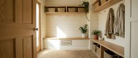 Bright farmhouse mudroom with wooden bench, wicker baskets, coat hooks, and potted plants, featuring shiplap walls and natural light.