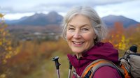 Senior Caucasian woman hiker smiling outdoors with trekking poles and backpack against autumn mountain landscape for active aging lifestyle content.