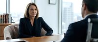 Professional middle-aged Caucasian businesswoman in black suit conducting serious meeting with male colleague in modern corporate office setting.